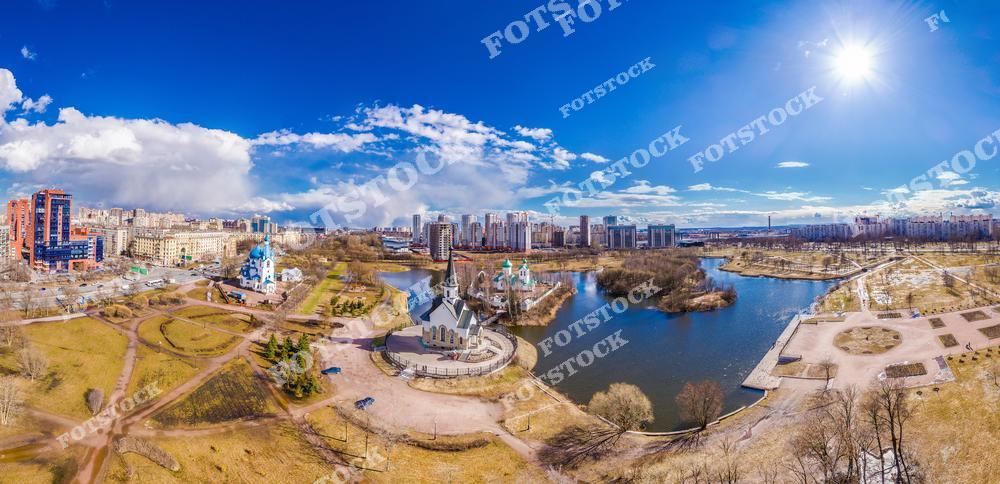 На снимке запечатлен Пулковский парк, расположенный в Санкт-Петербурге, с озером, отражающим ясное голубое небо. Вокруг парка видны современные жилые кварталы, гармонично вписанные в городской ландшафт. Весенний солнечный день подчеркивает красоту природы и уют этого места. Парк является популярным местом отдыха для горожан.